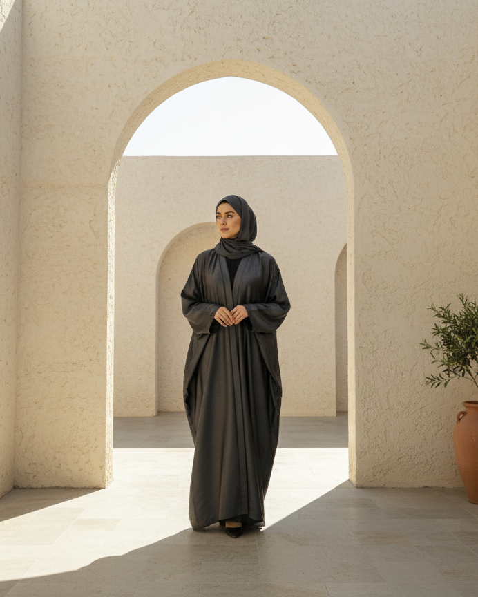 Woman in a black abaya standing in a sunlit archway with a potted plant nearby.
