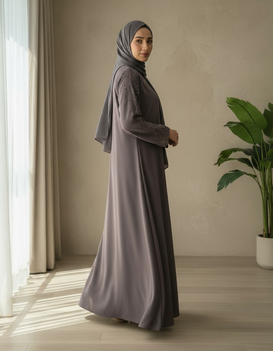 Woman in a long, dark gray dress and hijab standing in a room with a plant and window.