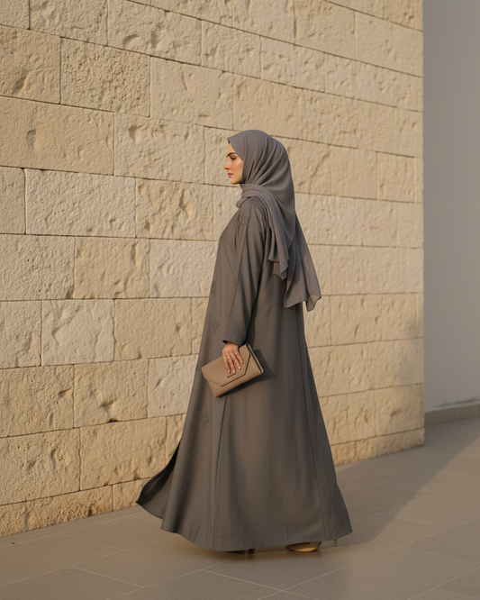 Woman in a long gray dress and hijab standing against a textured beige wall.