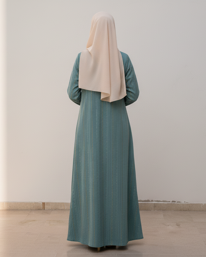 Person wearing a long teal dress and beige hijab standing in a room with a plant and white wall.