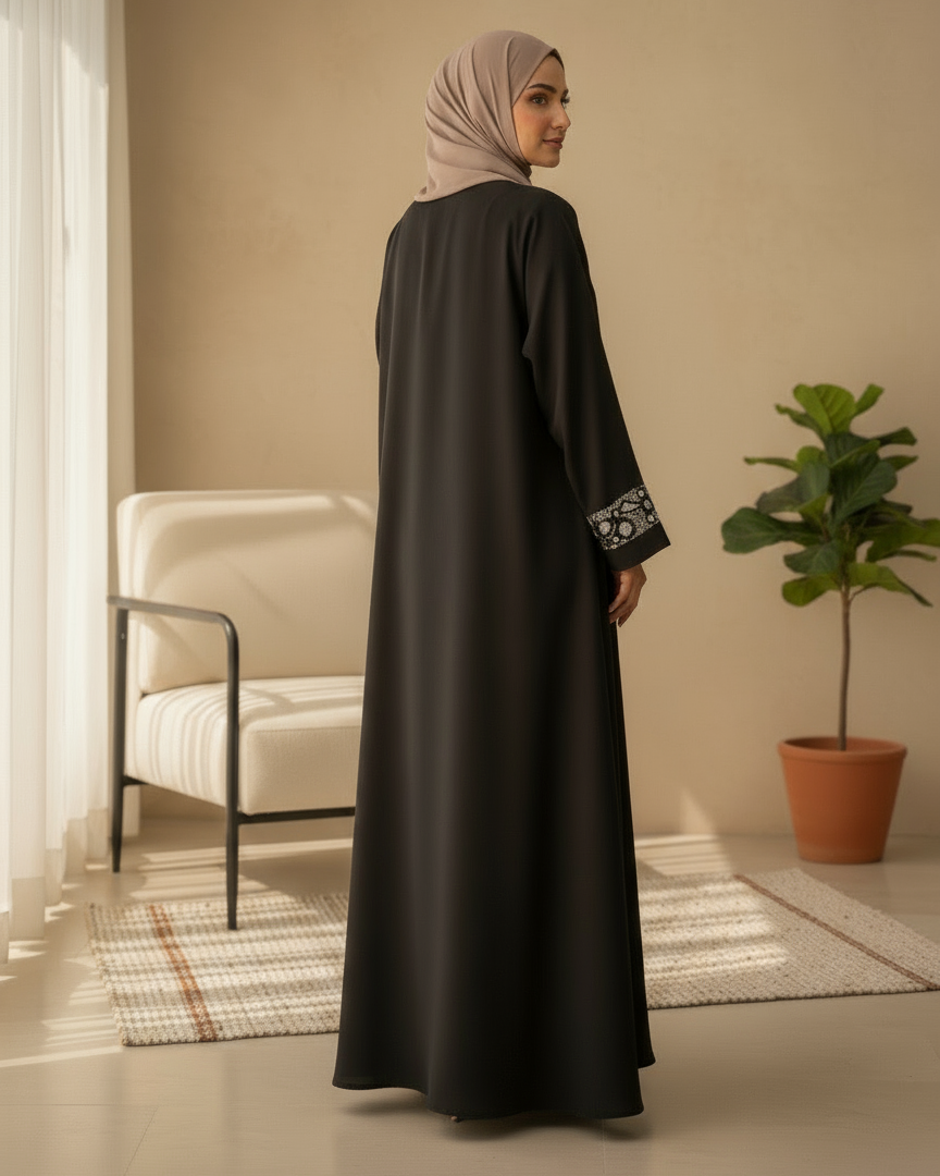 Woman in a black abaya with white embroidery standing in a room with a beige wall, white chair, and potted plant.