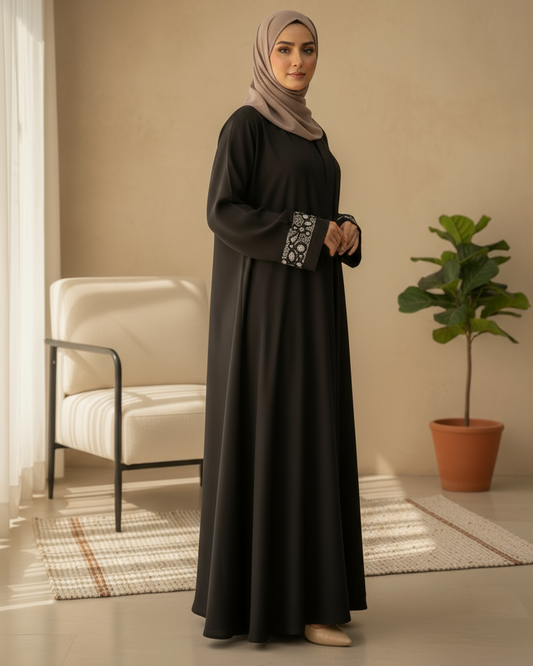 Woman wearing a black abaya with floral embroidery in a room with a beige wall, white chair, and potted plant.