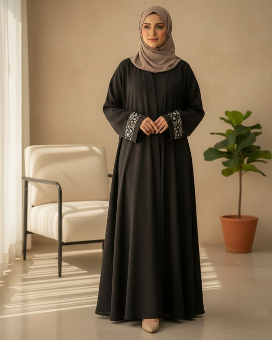 Woman wearing a black dress with patterned cuffs in a room with a beige wall, white chair, and potted plant.