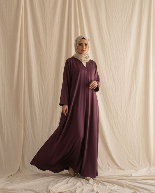 Woman in a long burgundy dress and hijab standing against a beige curtain.