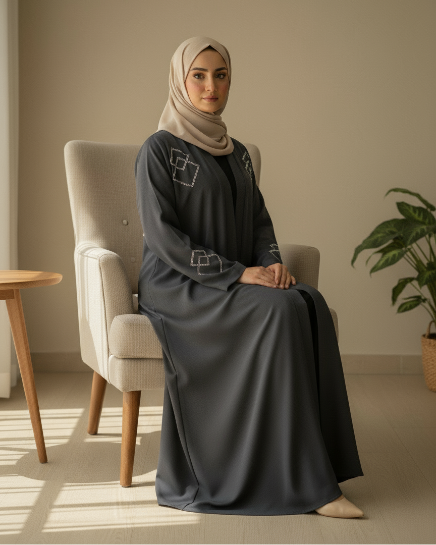 Woman in a gray abaya sitting on a chair in a well-lit room with a plant in the background.