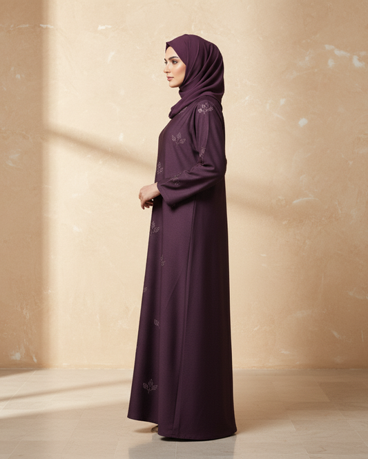 Woman in a long dark purple dress and hijab standing against a beige wall.