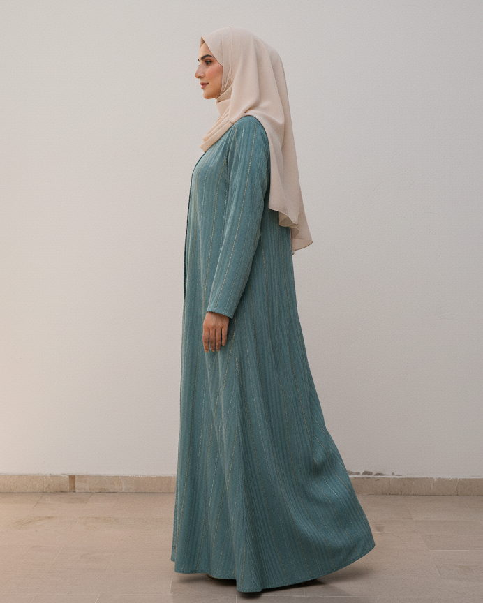 Woman wearing a long teal dress and beige hijab standing against a white wall.