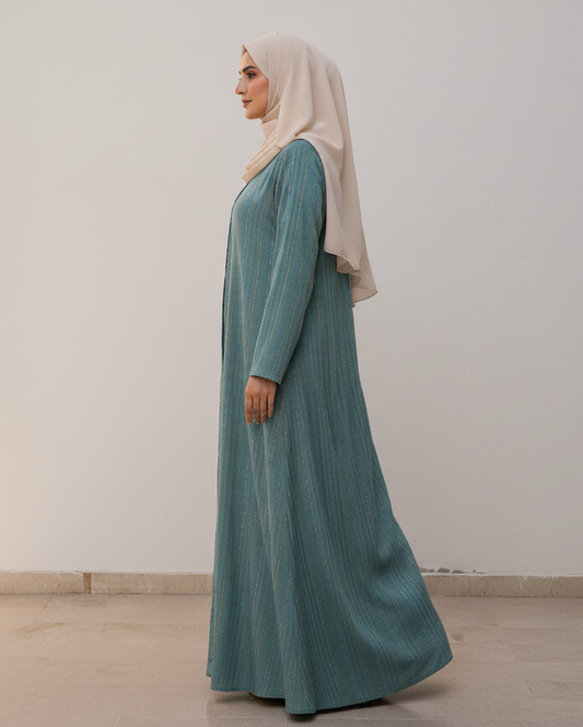 Woman wearing a long teal dress and beige hijab standing against a white wall.