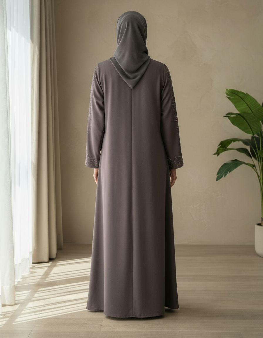 Person wearing a long, dark gray dress standing in a room with a plant and light streaming through curtains.