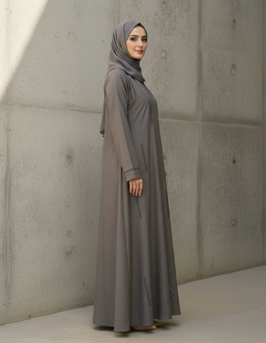 Woman wearing a long, light gray dress and hijab standing against a concrete wall.