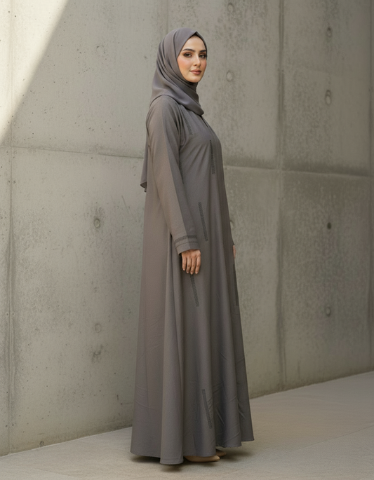 Woman wearing a long, light gray dress and hijab standing against a concrete wall.
