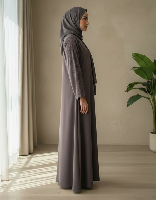 Woman in a long, dark gray dress standing in a room with a plant and window.