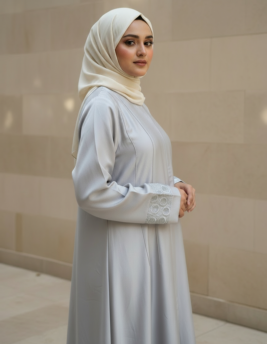 Woman wearing a light gray dress with lace details and a beige hijab against a beige wall.