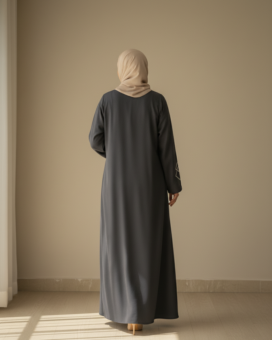 Person wearing a long dark dress and beige hijab standing in a room with a neutral wall.